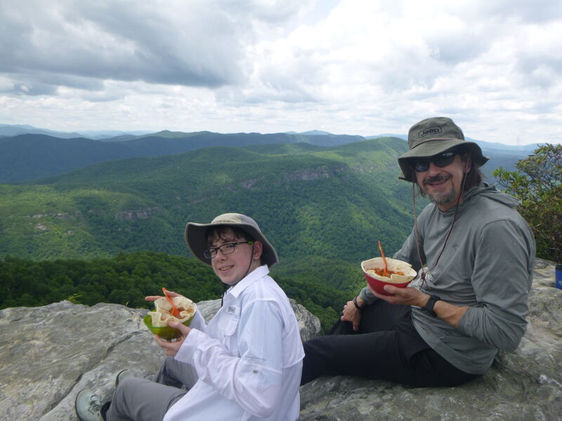 Two people are sitting on a rocky outcrop, enjoying a meal with a scenic mountain backdrop. The person on the left is younger, wearing glasses, a hat, and a white shirt, holding a bowl. The person on the right, presumably older, also wears a hat and is holding a bowl. They both appear to be on a hike, taking a break to eat and enjoy the view of the lush, green mountains under a cloudy sky.